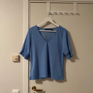 Blå v-ringad topp från Vero Moda - Snygg ljusblå topp från Vero Moda med v-ringning och korta ärmar med smockdetalj. Toppen har en lätt strukturerad yta och en avslappnad passform som känns både trendig och bekväm. Perfekt att styla med jeans eller kjol för en fräsch look. Katt finns i hemmet 