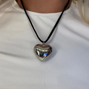 Halsband med silverfärgat hjärta - Snyggt halsband med ett stort, blankt hjärtformat hänge i silverfärg. Hänget sitter på ett svart band som ger en cool kontrast🌼