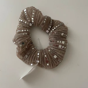 Glittrig beige scrunchie från Glitter - Superfin scrunchie i beige med glittrigt tyg och silvriga paljetter. Perfekt accessoar för att lyfta din frisyr med extra bling. Tjock och fluffig modell som ger volym till håret. Säljs med originaltag kvar. Aldrig använd och normal storlek!