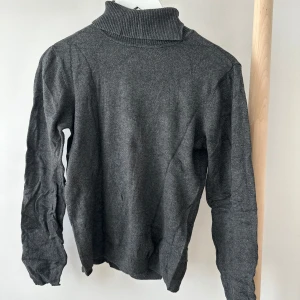 Mörkgrå polotröja från Vero Moda - Mörkgrå polotröja från Vero Moda i mjukt material. Tröjan har en hög ribbad krage och långa ärmar. Enkel och stilren design som passar perfekt till jeans eller kjol. Skön passform och lätt att matcha med andra plagg.