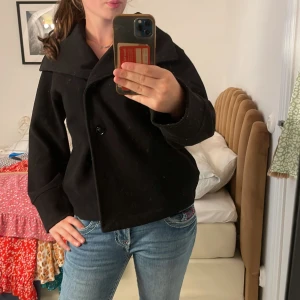 Intressekoll‼️ Svart skepparkavaj med bred krage - Säljer en svart skepparkavaj med bred krage och dubbelknäppning. Jackan har en boxig passform och är tillverkad i ett tjockare ullmaterial som håller dig varm under kyliga dagar. Perfekt för dig som gillar en klassisk och stilren look.