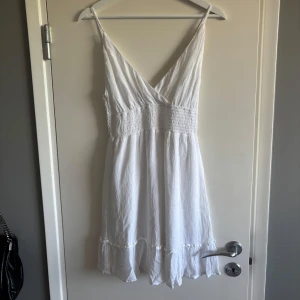 Vit somrig kortklänning med volang - Superfin vit kortklänning med smala axelband, v-ringning och smockad midja. Klänningen har volangdetaljer längst ner och är tillverkad i ett lätt, luftigt tyg som passar perfekt för varma dagar.