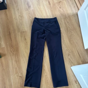 Low waist kostymbyxor bootcut - Snygga mörkblå kostymbyxor med klassisk bootcut och pressveck. Byxorna har hällor för bälte, snedställda fickor fram och fuskfickor bak. Perfekta för en clean och stilren look. Materialet känns som bomull eller polyester. Längs ner på byxorna har de spruckit lite men syns inte på, syns heller inte att de är mörkblå typ ❤️ skrivhjärna vid pris eller frågor 