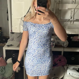 Blommig blå offshoulder kortklänning - Supersöt blå kortklänning med vita blommor och offshoulder-modell. Klänningen är figurnära och har korta ärmar. Materialet är ribbat och stretchigt, perfekt för sommarens varma dagar. Säljer då den inte kommer till användning längre!