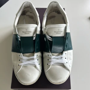 Valentino Garavani sneakers vit/grön - Snygga Valentino Garavani sneakers i vitt skinn med bred grön läderdetalj över snörningen. Klassisk rund tå, vita snören och ikoniska nitar baktill på sulan. Perfekta för dig som gillar exklusiva och stilrena sneakers.