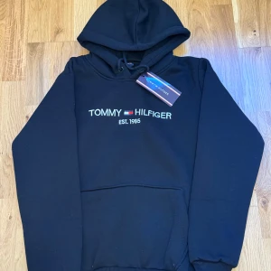 Svart hoodie Tommy Hilfiger - Svart hoodie från Tommy Hilfiger med broderad logga och text 'EST. 1985' på bröstet. Klassisk huva med snörning och stor magficka framtill. Mjuk bomullsblandning som är perfekt för chill dagar. Snygg och enkel stil som funkar året runt.