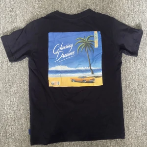 Svart t-shirt Chasing Dreams Only & Sons - Svart t-shirt från Only & Sons med trycket 'Chasing Dreams' på bröstet och en stor färgglad print på ryggen med strand, palm och bil. Klassisk passform och rund hals. Perfekt för dig som gillar streetstyle och snygg stl, Ny pris 249.