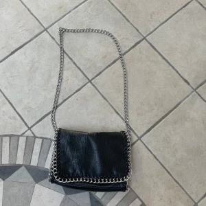 Svart axelväska med kedja från Tiamo - Cool svart axelväska från Tiamo i skinnimitation med grov silverfärgad kedja som axelrem och runt kanterna. Väskan har dragkedjefack både inuti och på baksidan samt magnetlås. 