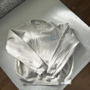 Beige sweatshirt Les Deux L - Beige sweatshirt från Les Deux i storlek L med blått tryck både fram och bak. Tröjan har rund halsringning, ribbade muddar och en relaxed passform. Materialet är mjukt och skönt, perfekt för chill dagar. Snyggt Harajuku-inspirerat print på ryggen.