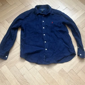 Marinblå linneskjorta Polo Ralph Lauren - Snygg marinblå skjorta från Polo Ralph Lauren i linne med klassisk krage och vita knappar. Skjortan har lång ärm och den ikoniska röda loggan broderad på bröstet. Perfekt för en stilren och avslappnad look.