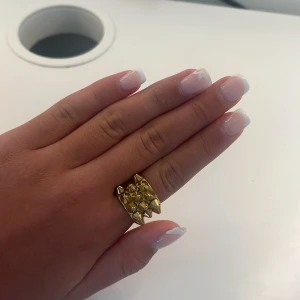 Guldig statementring med nitar - Cool och chunky ring i guldton med flera små konformade nitar på ovansidan. Ringen har en edgy vibe och passar perfekt för dig som vill sticka ut med accessoarerna. Metallisk finish och bred design gör den till ett riktigt statement.