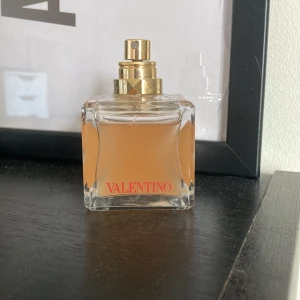 Valentino parfym i glasflaska - Elegant parfym från Valentino i en fyrkantig glasflaska med guldfärgad spraykork. Flaskan har en mjukt böjd design och parfymen inuti är ljus orange. Valentino-loggan syns tydligt i rött på framsidan. Obs sprutet är lite starkt!
