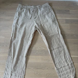Beige randiga linnebyxor med snörning - Jag säljer nu mina linnebyxor! Snygga beige linnebyxor med vita ränder och avslappnad passform. Byxorna har snörning i midjan och raka ben, perfekta för en skön dag. 
