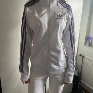 Vit Adidas ziphoodie med lila ränder - Vit ziphoodie från Adidas med klassiska lila ränder längs ärmarna och Adidas-logga på bröstet. Jackan har dragkedja framtill, hög krage och ribbade muddar. Perfekt för en sportig och avslappnad stil.
