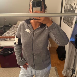 Grå zip-hoodie från Nike - Grå zip-hoodie från Nike med vit logga på bröstet och stor vit text på ena ärmen. Jackan har huva, hel dragkedja och är långärmad. Perfekt för chill dagar eller när du vill ha en sportig look.