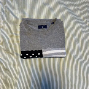 Grå stickad tröja från GANT - Snygg grå stickad tröja från GANT med amerikansk flagga i svart, vitt och grått på bröstet. Rund halsringning, ribbade muddar och lång ärm. Broderad text på ena ärmen. Perfekt för dig som gillar klassisk men ändå unik stil.