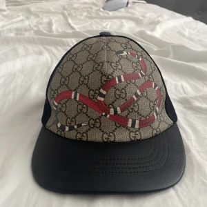 Gucci keps sällsynt ÄKTA !  - Snygg keps från Gucci med klassiskt GG-mönster i beige och svart, prydd med en röd orm framtill. Skärmen är i svart skinn och baksidan har svart mesh samt justerbart band i grönt och rött. Tillverkad i polyester, bomull och skinn. Perfekt statement-piece för dig som vill sticka ut.                