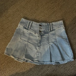 Ljusblå plisserad jeanskjol - Superfin ljusblå jeanskjol med plisserad design och låg midja. Kjolen har bred linning med tre silvriga knappar framtill och hällor för bälte. Snygg att styla med crop top eller hoodie.