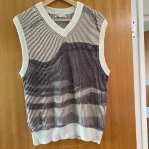 Stickad väst från Zara i beige och grå - Stickad väst från Zara med ribbad struktur och v-ringning. Västen har ett vågigt mönster i beige och grå toner, vilket ger en cool och modern look. På sista bilden kan man se lite skador den har men inget man ser på långt håll