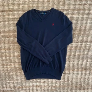 Marinblå v-ringad tröja Polo Ralph Lauren - Snygg marinblå v-ringad tröja från Polo Ralph Lauren med den klassiska röda loggan broderad på bröstet. Långärmad modell i mjuk bomull, perfekt för lager-på-lager och chill stil.