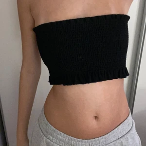 Svart bandeau tubtopp från Gina Tricot - Svart bandeau tubtopp från Gina Tricot i smockat tyg med volangkant nedtill. Toppen är axelbandslös och har en tight passform som sitter snyggt på kroppen. Perfekt till sommaren och enkel att matcha med både jeans och kjol.