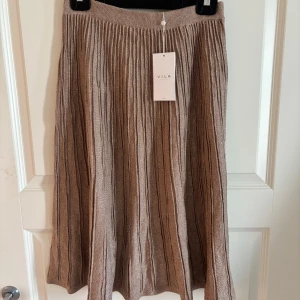 Glittrig beige långkjol från Vila - Supersnygg beige långkjol från Vila med glittriga inslag och ribbad struktur. Kjolen har ett mjukt, stickat material och elastisk midja för skön passform. Perfekt för dig som vill ha en chic och trendig look med lite extra shimmer. Nypris 450kr