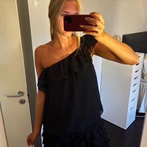 Svart oneshoulder topp med volang - Säljer en svart oneshoulder topp med volangdetalj längs halsringningen.