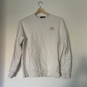 Vit sweatshirt från Dolce & Gabbana - Snygg vit sweatshirt från Dolce & Gabbana med diskret logopatch på bröstet. Tröjan har rund hals, långa ärmar och ribbade muddar. Tillverkad i mjuk bomull för en clean och stilren look. Perfekt för dig som gillar exklusiva basics. Storlek: S (herr) 