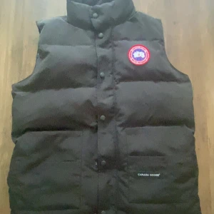 Svart dunväst från Canada Goose - Svart dunväst från Canada Goose med klassisk logga på bröstet. Västen har hög krage, tryckknappar framtill och två fickor med broderad logotyp. Perfekt för lager-på-lager och riktigt varm tack vare dunfyllningen. Västen är nästan helt ny jag bara haft på mig den 6-7 gånger. 