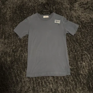 Blå t-shirt från Stone Island  - Säljer en stilren blå t-shirt från Stone Island Junior med vit logga tryckt på bröstet. T-shirten har rund halsringning och klassisk passform. Perfekt för dig som vill ha något enkelt men snyggt.