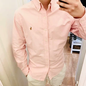 Ljusrosa Skjorta från Polo Ralph Lauren (Slim Fit) - Säljer en ljusrosa skjorta från Polo Ralph Lauren med klassisk button-down krage och broderad logga på bröstet. Skjortan har långa ärmar och knäppning framtill. Perfekt för en stilren och fräsch look. Det står att skjortan är storlek L men det är det inte, skjortan är Small passar XS också Slim fit. Snabb Leverans!