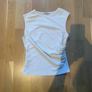 Vit ribbad ärmlös topp från H&M - Säljer en vit ärmlös topp från H&M i storlek XS. Toppen är ribbad och har en lätt rynkad detalj på ena sidan vilket ger en snygg passform. Perfekt för varma dagar och enkel att matcha med olika outfits.