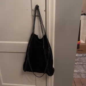Svart axelväska med kedjedetaljer - Säljer en supersnygg svart trendig axelväska med kraftiga silverfärgade kedjor som axelremmar och längs kanterna. Väskan har en mjuk form och innerfack med dragkedja🥰. Perfekt för dig som vill ha en stilren och rymlig och trendig accessoar💞💞