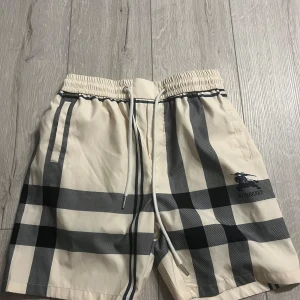 Beige och svart rutiga shorts från Burberry - Snygga beige shorts med klassiskt svart och grått rutmönster från Burberry. Shortsen har resår i midjan med vit snörning och är perfekta för sommaren. Tillverkade i Italien och har en stilren look med Burberry-logga på benet. Pris kan diskuteras!