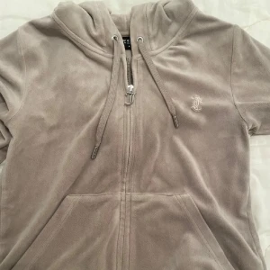 Beige velour hoodie från Juicy Couture - Mysig beige hoodie i velour från Juicy Couture med dragkedja framtill, huva med dragsko och fickor. Liten JC-logga broderad på bröstet. Perfekt för en avslappnad och trendig look.