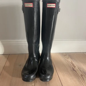 Svarta höga gummistövlar från Hunter - Klassiska svarta gummistövlar från Hunter med högt skaft och rund tå. Perfekta för regniga dagar och har diskret spänndetalj på sidan. Hunter-logga framtill upptill på skaftet.