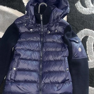 Blå dunväst med stickade ärmar från Moncler - Snygg blå dunväst från Moncler med stickade svarta ärmar och huva. Inga defekter helt ny!
