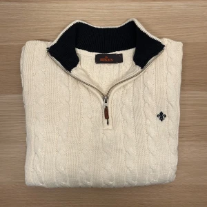 Morris Kabelstickad Half-Zip Creme S/XS - Säljer en klassisk vit kabelstickad tröja från Morris med halv dragkedja och mörk kontrastkrage. Tröjan har lång ärm, ribbade muddar och en broderad logga på bröstet. Perfekt för en stilren och tidlös look. Passar S-XS