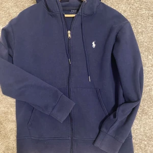 Mörkblå kofta från Polo Ralph Lauren - Säljer en mörkblå hoodie från Polo Ralph Lauren med dragkedja. Tröjan har två fickor framtill och en broderad vit logga på bröstet. Enkel att matcha. Är i bra skick. Säljer då jag inte använder längre