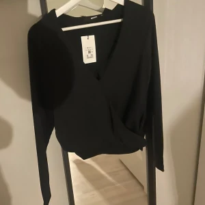 Svart omlottopp från Bik Bok - Säljer en svart omlottopp från Bik Bok med lång ärm och djup v-ringning. Toppen har en elegant omlottdesign som ger en snygg siluett och passar perfekt till många olika outfits.