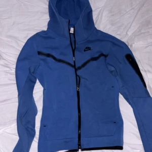 Blå zip-hoodie från Nike - Säljer en blå hoodie från Nike med dragkedja och huva. Jackan har svarta detaljer, inklusive en sned dragkedja över bröstet och Nike-logga. Fickor framtill och ribbade muddar. Perfekt för en sportig stil.