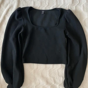 Svart croppad topp från SHEIN - Säljer en svart croppad topp från SHEIN med långa puffärmar och fyrkantig ringning. Toppen är i ett mjukt, syntetiskt material och har en tight passform. Perfekt att styla med höga jeans eller kjol.