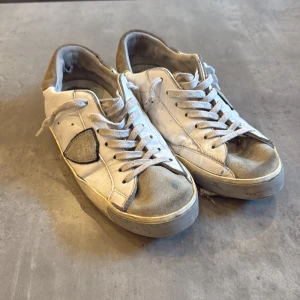 philippe model - Säljer ett par vita sneakers med grå detaljer och beige häl. Skorna har snörning och platt sula, samt rund tå. Perfekta för dig som gillar en klassisk och enkel stil med en touch av kontrastfärg.
