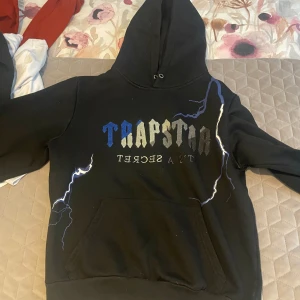 Svart hoodie från Trapstar S  - Säljer en svart hoodie från Trapstar med tryckt text och blixtdetaljer i blått och vitt. Tröjan har huva och magficka, samt ribbade muddar vid ärmslut och nederkant. Perfekt för en streetwear-look.