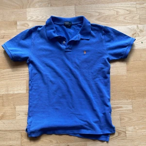 Blå pikétröja från Ralph Lauren - Klassisk blå pikétröja från Ralph Lauren med liten broderad logga på bröstet. Tröjan har korta ärmar, krage och knäppning med två knappar. Perfekt för en stilren och avslappnad look. Storleken är L men är liten i storlek så den egentliga storleken är M, den enda defekten är 2 röda sträck på baksidan därav priset