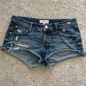 Lågmidjade shorts - Säljer ett par blå jeansshorts från Just Jeans med slitna detaljer och råa kanter. Klassisk femficksmodell med knapp och dragkedja framtill. Perfekta för varma dagar och en avslappnad stil.