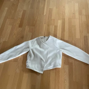 Vit omlott kappa - Säljer en stilren creme vit kappa med omlott-design. Kappan har lång ärm och en minimalistisk look som passar perfekt till många outfits. Enkel och modern passform med snyggt snitt framtill.