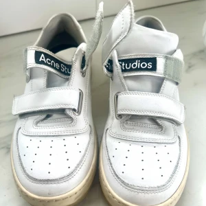 Vita sneakers med kardborre från Acne Studios storlek 39  - Säljer ett par vita sneakers från Acne Studios med två breda kardborreband och perforerad tå. Skorna har en beige sula och en ikonisk smiley-detalj bak. Perfekta för en clean och stilren look.   Använda vid tre tillfällen så väldigt bra skick! 