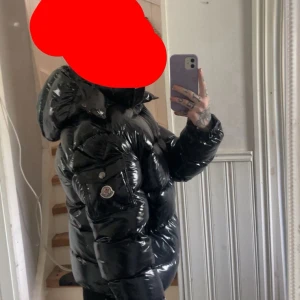 Svart glansig dunjacka från Moncler - Säljer en svart, glansig pufferjacka från Moncler med huva och dragkedja. Jackan har en quiltad design och Moncler-logga på ärmen. Jag säljer den då jag inte tycker den passar min stil använd fåtal gånger köptes i vintras ny inga skador eller andra konstigheter på jackan storlek 2 (m)