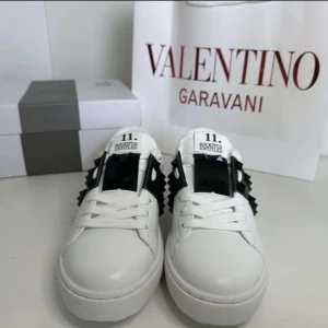 Valentino Garavani vita sneakers med svarta nitar - Säljer ett par vita Valentino Garavani sneakers med svart band och svarta nitar på sidorna. Skorna har rund tå, snörning och en platt sula. Perfekta för dig som vill ha en lyxig och edgy look.